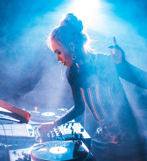 concentrated dj woman with blonde hair gesturing and using dj equipment in nightclub with smoke