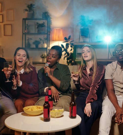 Group of young intercultural friends singing karaoke while sitting on couch